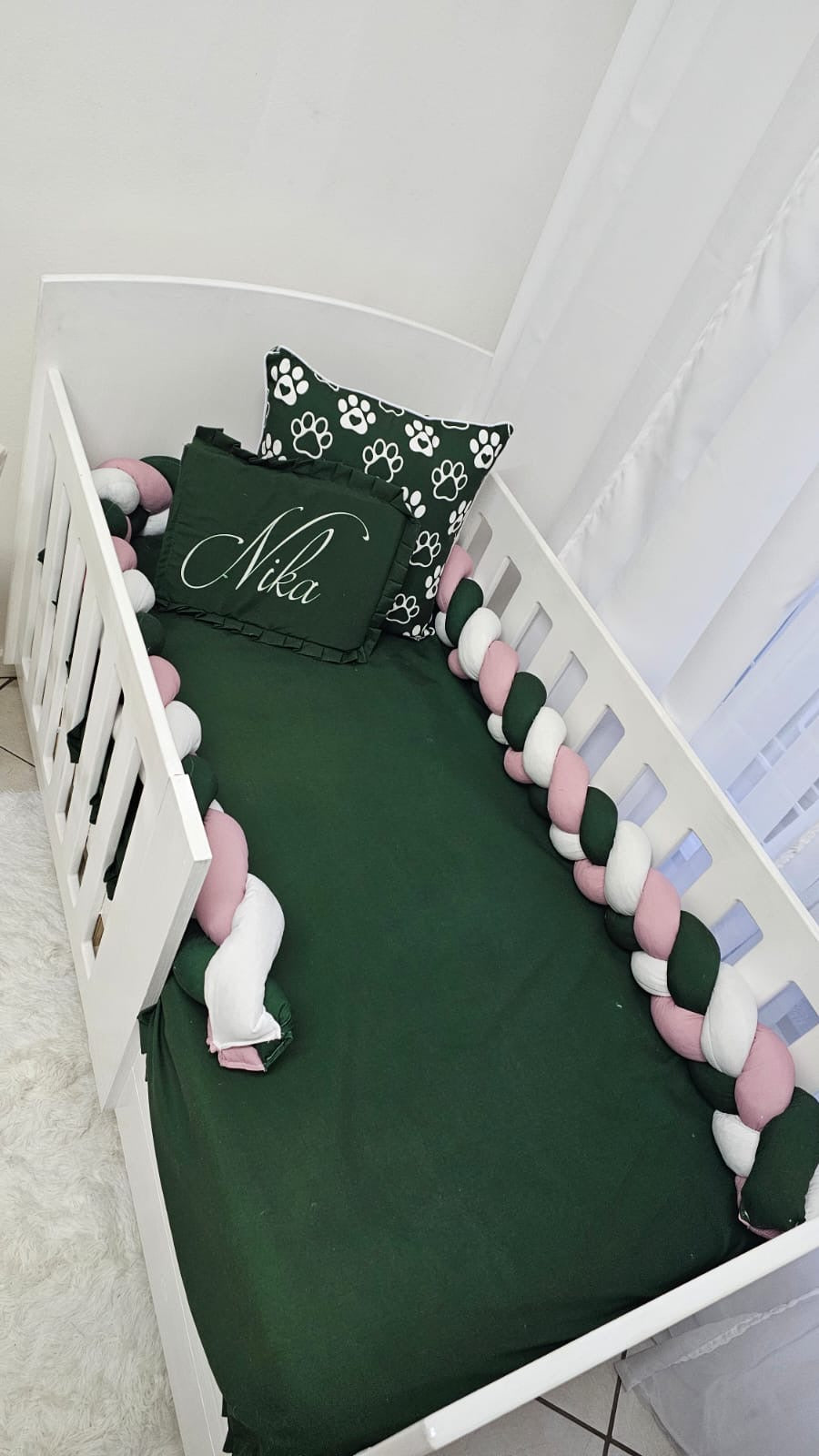 Children's crib with green bedding, pink and white bumper, and decorative pillow.