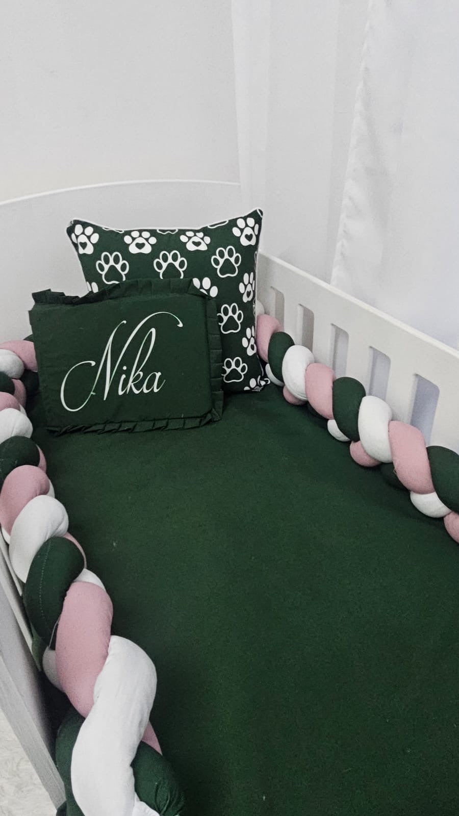 Crib with green bedding, pink and white bumper, and decorative pillows with paw prints and 'Nika' text.