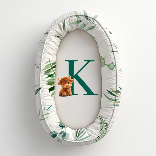 Brave and Bold baby nest featuring a sweet Highland cow, personalised green initial “K” and soft botanical green leaf print on a clean white background | Brave and Bold babanes met ’n oulike Highland-bees, gepersonaliseerde groen voorletter “K” en ’n sagte botaniese groen blaarpatroon op ’n skoon wit agtergrond.
