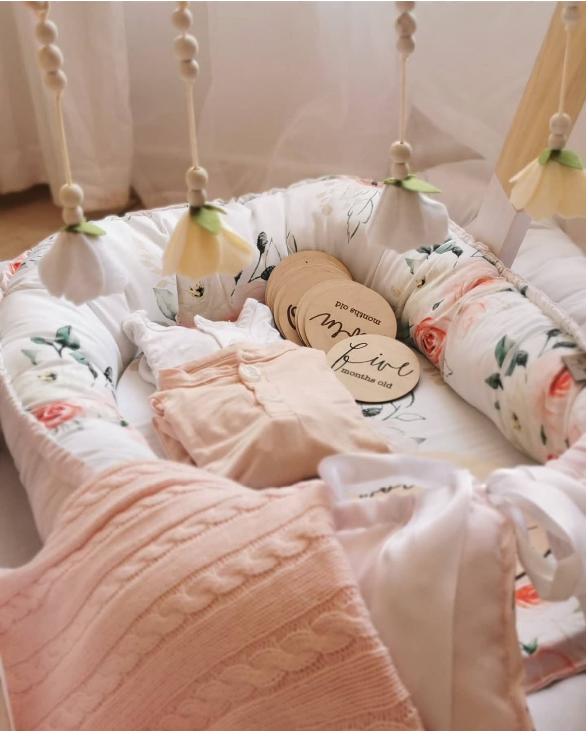 Custom baby nest featuring a soft floral print, blush pink knitted blanket, baby clothing and wooden milestone discs in a cosy nursery setting | Pasgemaakte babanes met ’n sagte blomdruk, ligpienk gebreide kombers, babaklere en hout-mylpaalskyfies in ’n knus babakamer-opset.