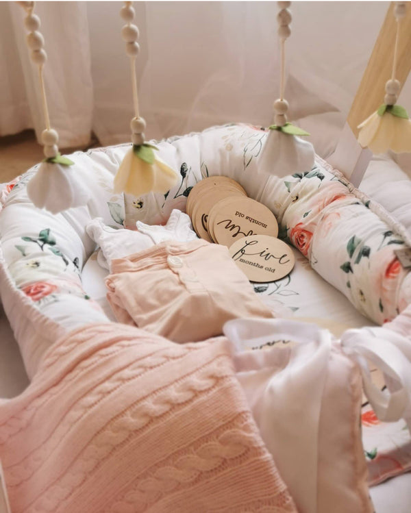 Custom baby nest featuring a soft floral print, blush pink knitted blanket, baby clothing and wooden milestone discs in a cosy nursery setting | Pasgemaakte babanes met ’n sagte blomdruk, ligpienk gebreide kombers, babaklere en hout-mylpaalskyfies in ’n knus babakamer-opset.
