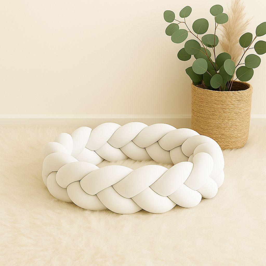 White braided cot bumper on a beige surface with a plant in the background