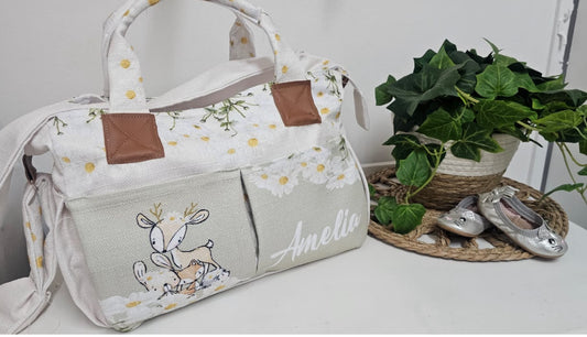 Personalised Daisy Woodlands nappy bag featuring a sweet deer, bunny and soft daisy details in neutral cream and sage tones, finished with front pockets and the name “Amelia” | Gepersonaliseerde Daisy Woodlands doeksak met ’n oulike takbok, hasie en sagte madeliefie-detail in neutrale room- en saliegroen kleure, afgewerk met voorsakke en die naam “Amelia”.