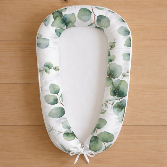 Eucalyptus Dreams baby nest featuring watercolour green eucalyptus leaves on white fabric, photographed flat lay on wooden surface
