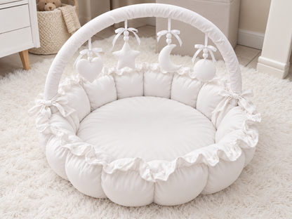 A white padded baby play mat with a ruffled border, a curved support arch, and hanging baby toys including a heart, star, and crescent moon, shown on a white rug in a nursery setting.