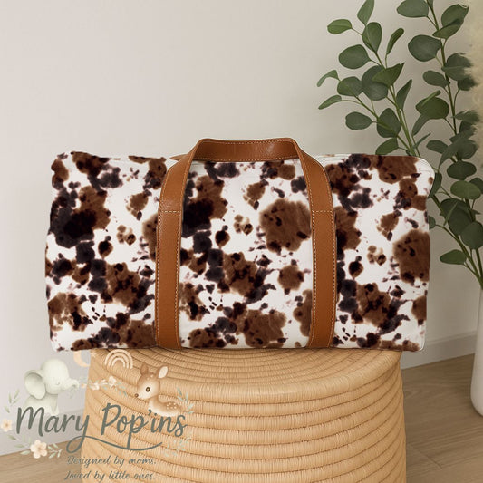 Highlands Trail hospital bag featuring a rustic brown, black and cream cowhide-inspired print with tan handles in a warm countryside style | Highlands Trail hospitaaltas met ’n rustieke bruin, swart en roomkleurige beesvel-geïnspireerde patroon met bruin handvatsels in ’n warm plattelandse styl.