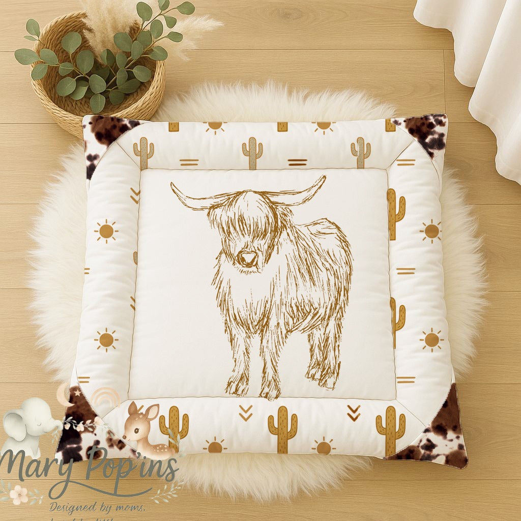 Highlands Trail baby playmat featuring a hand-drawn Highland cow with warm mustard cactus, sun and geometric details, finished with cowhide-style corner accents | Highlands Trail baba-speelmat met ’n handgetekende Highland-bees, warm mosterdkleurige kaktusse, son- en geometriese detail, afgewerk met beesvel-styl hoekaksente.