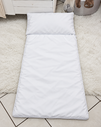 A white convertible toddler bed with an attached pillow, laid out on a white shag rug over a tiled floor.