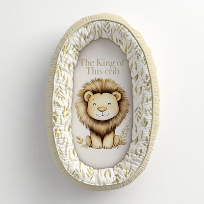Oat So Neutral Safari baby nest featuring a sweet lion with the phrase “The King of This Crib,” surrounded by soft olive leafy details in warm oat, cream and neutral tones | Oat So Neutral Safari babanes met ’n oulike leeu en die frase “The King of This Crib,” omring deur sagte olyfgroen blaardetail in warm hawer-, room- en neutrale kleure.