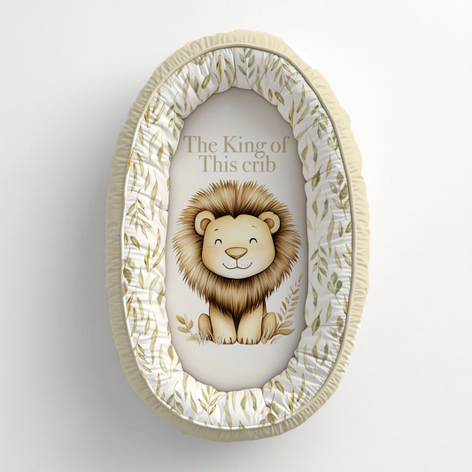 Oat So Neutral Safari baby nest featuring a sweet lion with the phrase “The King of This Crib,” surrounded by soft olive leafy details in warm oat, cream and neutral tones | Oat So Neutral Safari babanes met ’n oulike leeu en die frase “The King of This Crib,” omring deur sagte olyfgroen blaardetail in warm hawer-, room- en neutrale kleure.