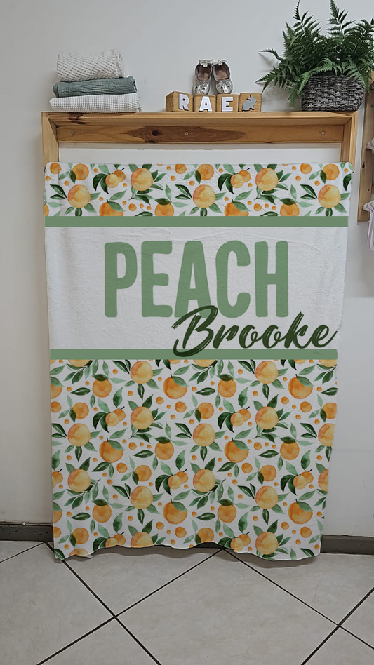 Peachy fleece blanket featuring a fresh peach fruit pattern with green leaves on a white background in soft warm nursery tones | Peachy fleece-kombers met ’n vars perskevrug-patroon en groen blare op ’n wit agtergrond in sagte warm kinderkamer-kleure.