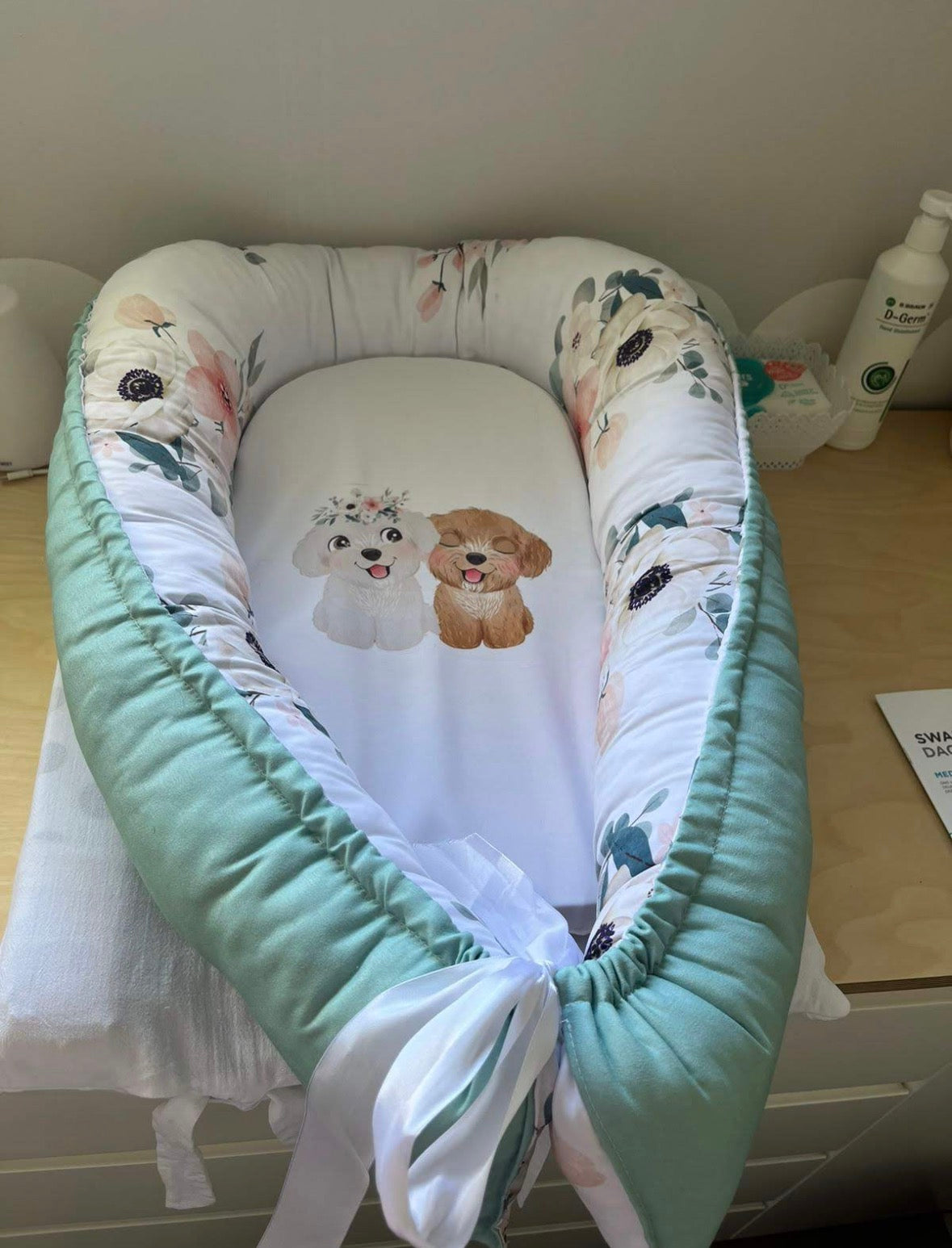 Rainbow Floral baby nest featuring sage green fabric with delicate pink and white flowers, photographed flat lay on wooden surface
