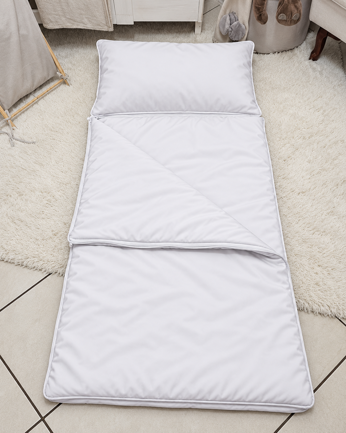 A white foldable sleeping pad with a matching pillow, laid out on a light-colored rug over a tiled floor.